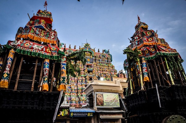 Two wooden chariots belonging to Nellaiappar temple stand before the temple Gopuram. The chariots are fully decorated with festive-looking cloth and flower garlands. 