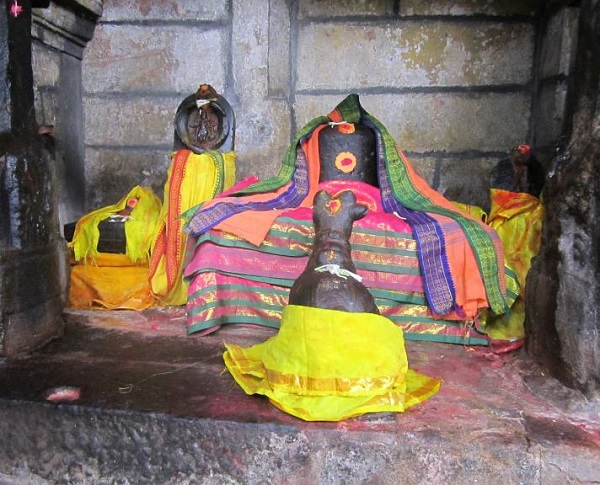 A stone shrine featuring a Lingam and a Nandi seated before it. The Lingam is covered with colourful silk materials and has a sandalwood and kunkumam pottu.