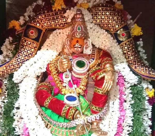 Pittapurathi Amman in a seated pose wearing a red, green and gold silk saree, bedecked with jewellery, flower garlands and a gold decoration.