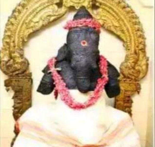 Lord Ganesha seated in front of a bronze decoration wearing a white dhoti and a garland and crown of pink flowers in Pittapurathi amman temple tirunelveli