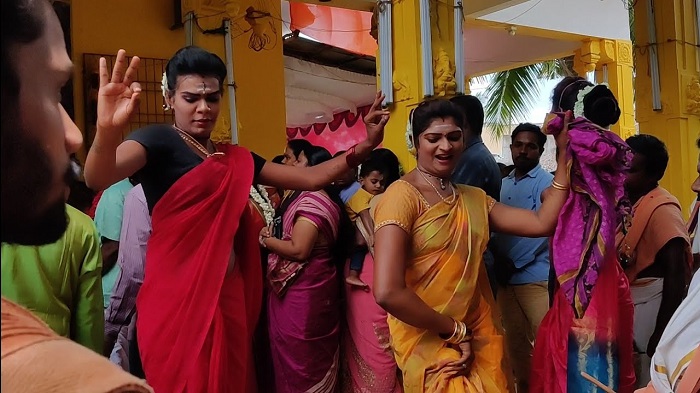 Folk artistes dressed as women performing Kaniyankoothu in a temple with devotees offering prayers at the temple visible in the background