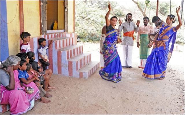 Two folk artistes dressed as women performing Kaniyankoothu with a drummer,and singers in the background and an audience of kids watching them perform in front of a temple