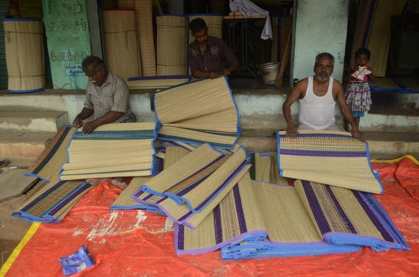 Three Pathamadai mat weavers working on Pathamadai mats, with rolls of mats neatly stacked and displayed in the background.
