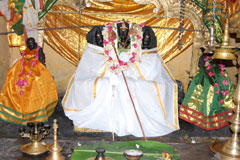 Sri Mayilerumperumal Saastha adorned in white with his consorts Sripoorna and Sri Pushkala adorned in yellow and green sarees, respectively. In front of them is a banana leaf with food offerings, and a brass bell and lamp stand.