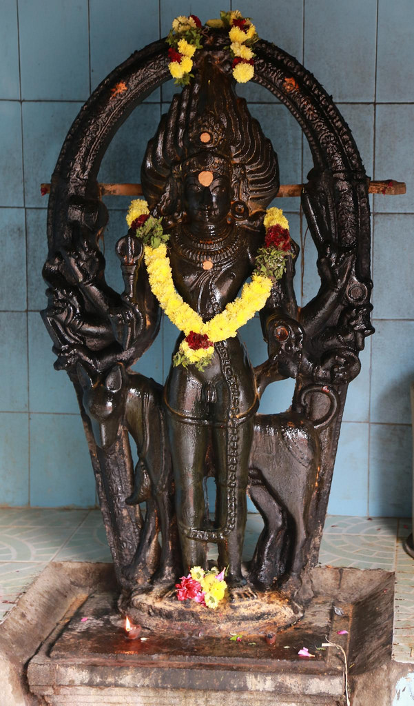 Statue of Lord Bairavar decked in a yellow flower garland and standing atop a stone pedestal with his dog.