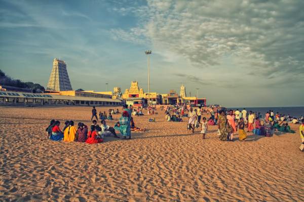 View of the Tiruchendur temple and the adjoining sea shore, the majestic temple gopuram against the blue sky, and the devotees sitting on the beach.