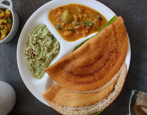 Picture of two crispy, golden brown wheat dosais placed on a banana leaf and arranged on a multi-cavity plate that holds a serving of sambar and coconut chutney.